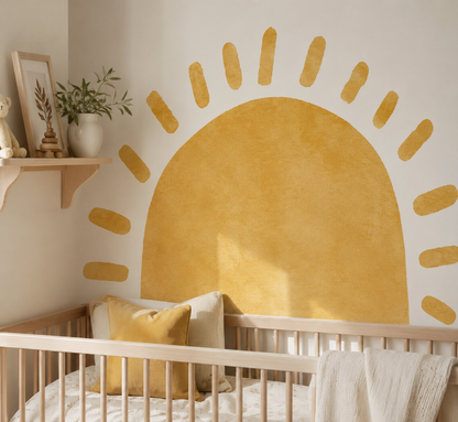 A styled nursery corner featuring a large textured mustard yellow sun wall decal above a light wood crib. The room has a warm off-white wall, a small wooden shelf with a vase of greenery and a framed print, and a white textured throw blanket draped over the crib.
